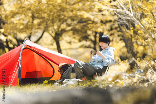 若い日本人女性がテントを張り、外に座りコーヒーを飲んでいる。/ソロキャンプ