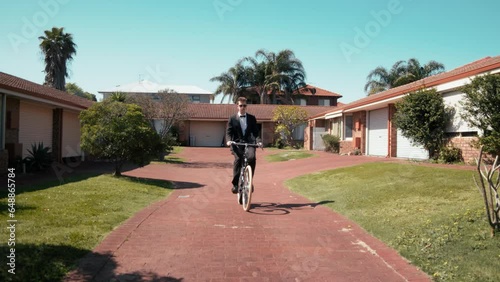 Well-dressed gentleman clad in a sleek black business suit or tuxedo and sunnies enjoys ride on a bicycle in a driveway of his house. photo