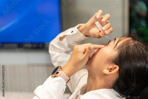 Young female Asian employee fingers her eyes and applies artificial tears or eye drops to the eyeballs to reduce eye strain while sitting at a desk in the company.