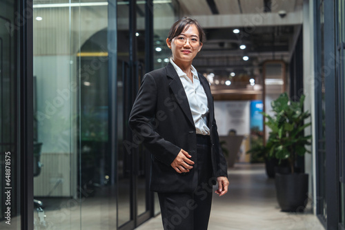 Young Asian woman wearing glasses, wearing a black suit, professional executive, standing confident in work success, standing in front of a company conference room.