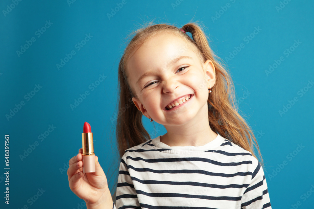 Happy little girl puts lipstick on her lips on blue background. Little trendy. little girl applying makeup Happy little girl puts lipstick on her lips on blue background. Little trendy. little girl applying makeup