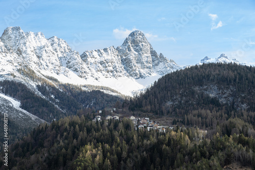 View of Cibiana di Cadore town at the foot of snow peaked mountains. Blue sky in a sunny winter day; Dolomites, Italian alps, Italy