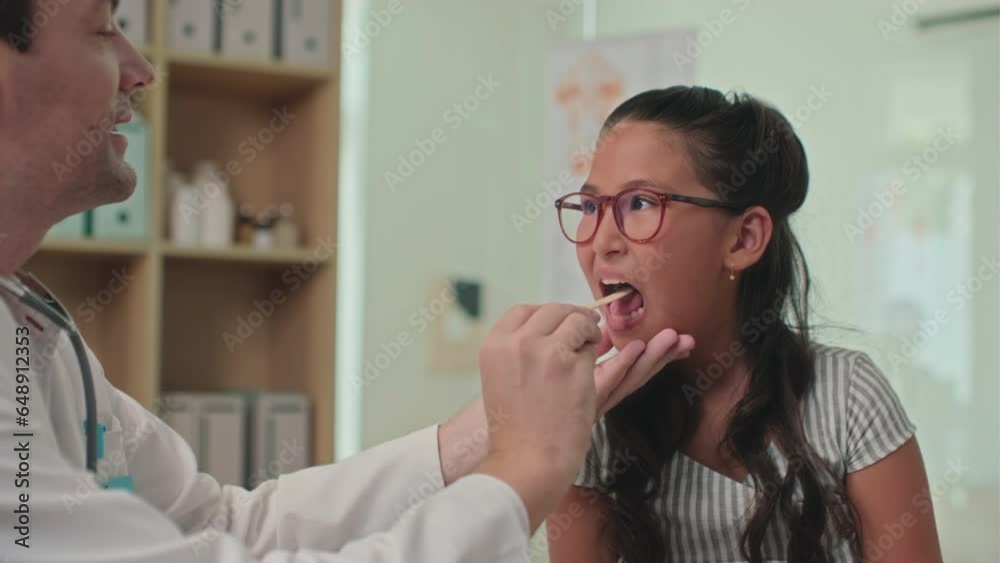 Medium shot of doctor using spatula when examining sore throat of teenage girl during visit at clinic Medium shot of doctor using spatula when examining sore throat of teenage girl during visit at clinic