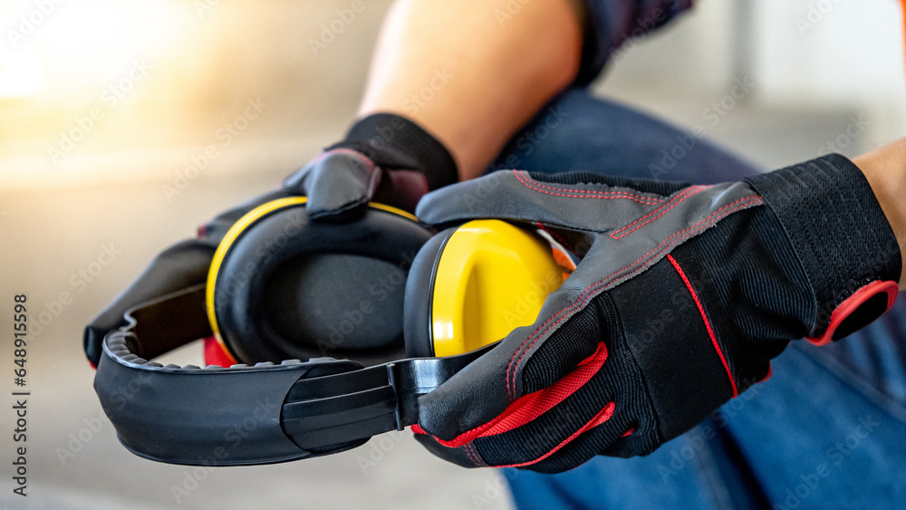 Male worker hand with black and red protective gloves holding yellow safety ear muffs or ear protectors preparing to wear on his head. Equipment for high noise reduction - obrazy, fototapety, plakaty 