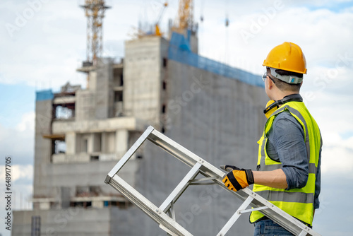 Asian man construction worker with green reflective vest, yellow safety helmet and ear muffs carrying aluminum ladder looking at unfinished building and tower crane at construction site