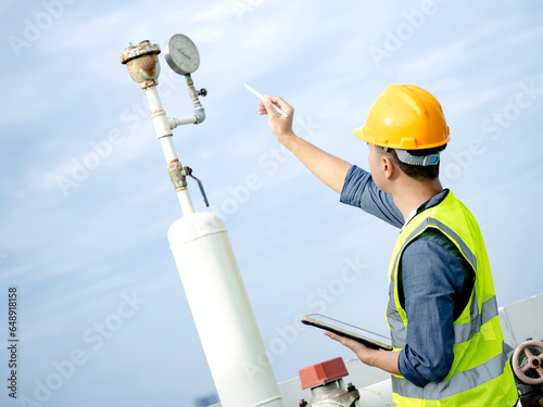 Male building inspector or maintenance worker man with green reflective vest, safety helmet using digital tablet and pen checking water pumping system pressure gauge on top of building photo