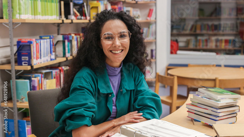 Portrait of a charming smiling Oriental girl with curly hair wearing glasses sitting at a school desk in a classroom or school library, backdrop of bookshelves smiling directly looking at the camera.
