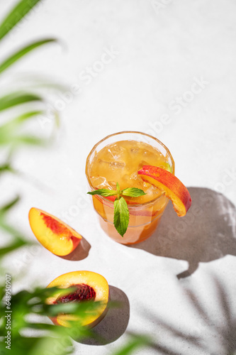 Refreshing peach tea with ice and mint. Homemade cold healthy vegetarian drink on a light background with fresh fruits, palm leaf and shadows.