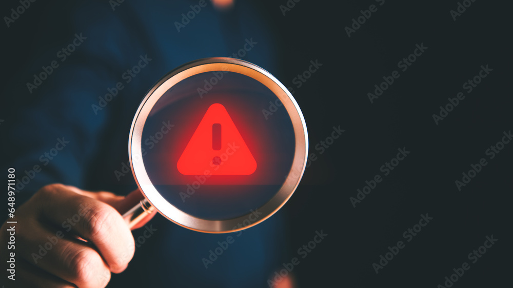 Red warning icon on a magnifying glass lens held by the businessman hand. - obrazy, fototapety, plakaty 
