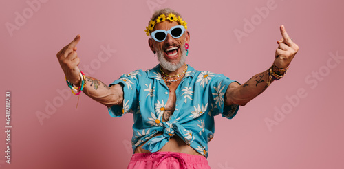 Cool senior man in stylish shirt and floral wreath gesturing and smiling against pink background