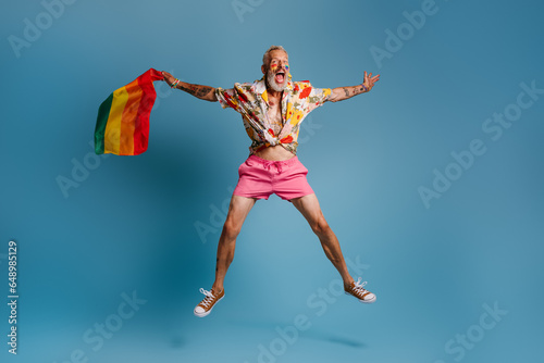 Full length of mature gay man carrying rainbow flag and smiling while jumping against blue background