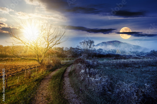 rural landscape with country road to mountain through agricultural fields behind fence and few trees in late autumn foggy weather with sun and moon at twilight. day and night time change concept