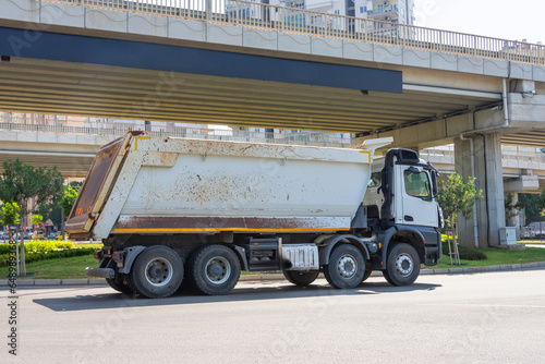 White dump truck with a scratched body drives along a city street under a bridge road.