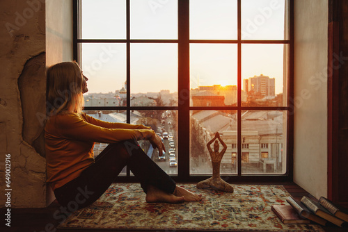 A woman sits on the windowsill and dreamily looks out of the window against the background of the city and sunset. The concept of harmony and balance in the city.