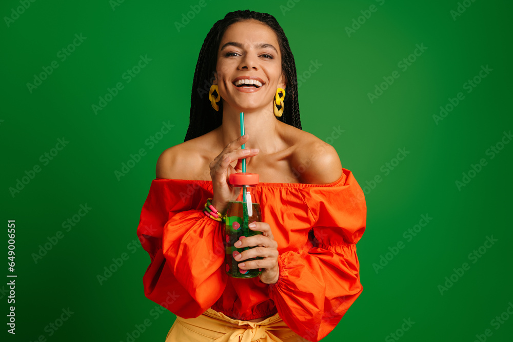 Joyful young woman holding bottle with lemonade and looking at camera against green background - obrazy, fototapety, plakaty 