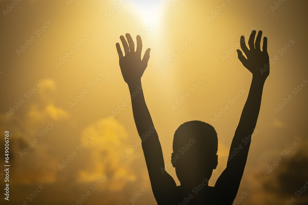 Silhouette of a young child, hands raised in prayer, his innocence illuminated by the soft light of the setting sun.