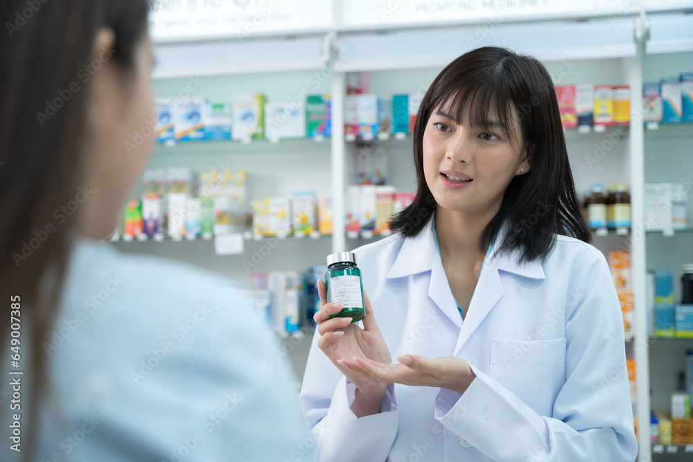 Young Asian pharmacist talking with customer at pharmacy counter. She tells customers about drug information at the pharmacy.