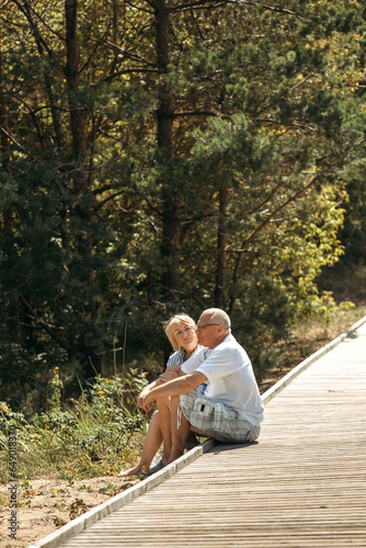 A happy elderly couple is sitting on a wooden path in a pine forest in the dunes and talking photo