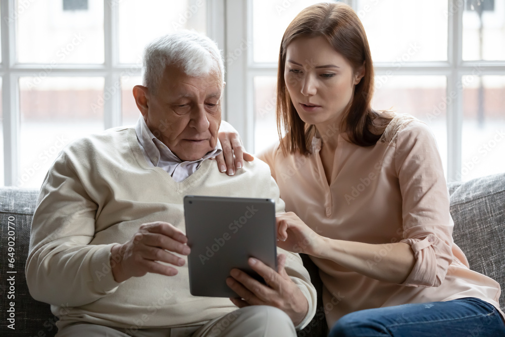 Focused adult girl teach elderly 80s father using modern pad gadget, browsing internet or news, smart old mature dad and grownup daughter surfing tablet device together, relaxing on couch at home
