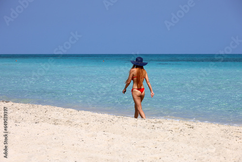 Woman in red bikini and sun hat walking by the sand on blue sea water background. Beach vacation on sunny coast
