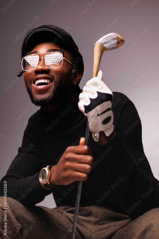 Portrait of smiling and elegant african american man in eyeglasses holding golf club isolated on grey - obrazy, fototapety, plakaty 