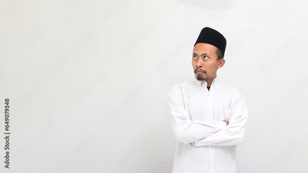an Angry young Indonesian man wearing a songkok or peci or kopiah, keeps his arms folded, looking aside at empty copy space with an annoyed and disapproving expression, isolated on a white background an Angry young Indonesian man wearing a songkok or peci or kopiah, keeps his arms folded, looking aside at empty copy space with an annoyed and disapproving expression, isolated on a white background
