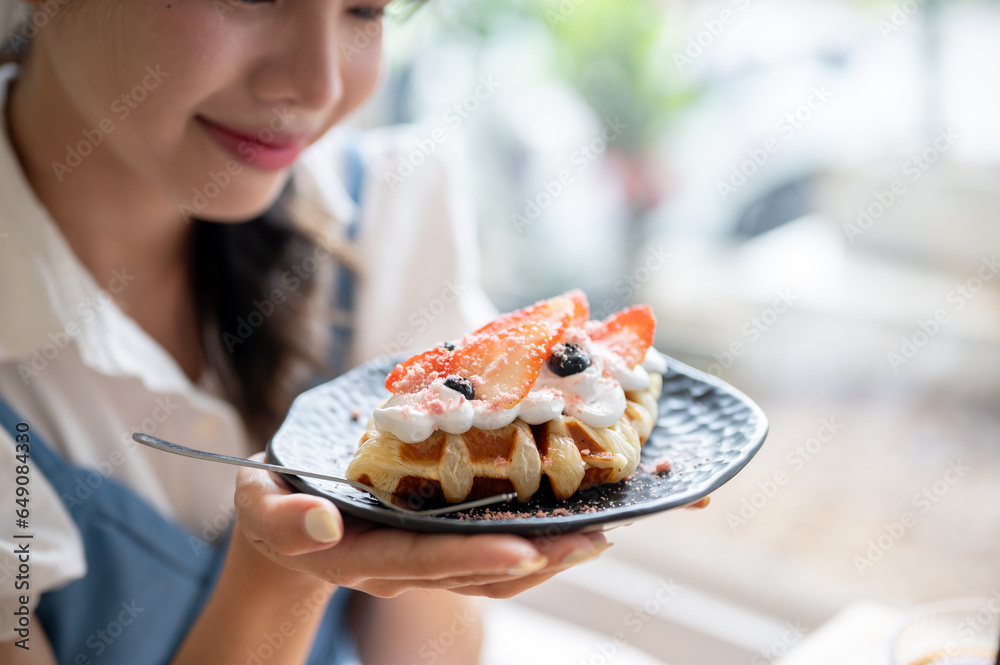 A happy and pretty Asian woman holding a plate of a yummy croissant waffle at a table in a cafe.