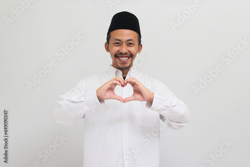 Smiling Young Indonesian man, wearing a songkok, peci, or kopiah, shows a love gesture with his hand, isolated on white background