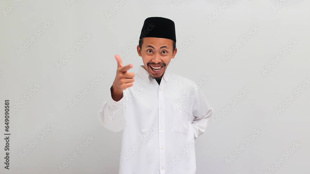 Excited Young Indonesian man, wearing a songkok, peci, or kopiah, is smiling and pointing his finger at the camera, choosing or picking, and inviting YOU to join, isolated on white background