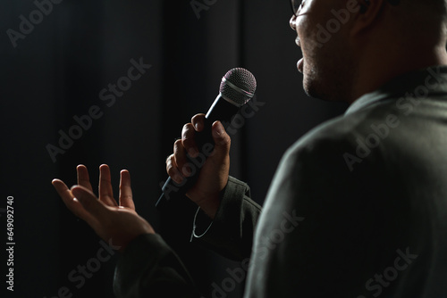 Confident successful speaker man talking on stage with spotlight strike through the darkness at corporate business event. Public speaker giving talk at conference hall. Stand up comedian.