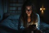 Dramatic Portrait of Single sad teen holding a mobile phone lamenting sitting on the bed in her bedroom with a dark light in the background