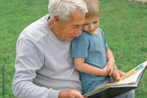 Happy grandfather reading book to curious grandson outdoors. Close up. Grandpa with grandchild spending time together. Family time comes in various forms. 4s year boy and senior man read story in park photo