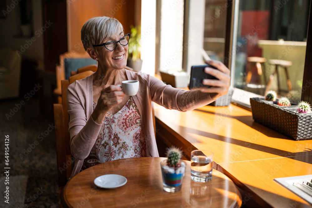Mature woman is drinking coffee in cafe. She is taking selfie. Mature woman is drinking coffee in cafe. She is taking selfie.