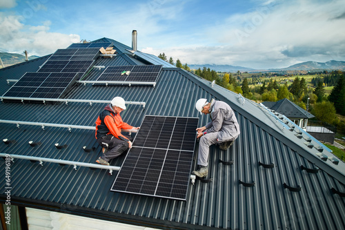 Roofers building photovoltaic solar module station on roof of house. Men electricians in helmets installing solar panel system outdoors. Concept of alternative and renewable energy. Aerial view.