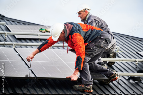 Builders installing photovoltaic solar panels on roof of house. Men engineers in helmets building solar module system with help of hex key. Concept of alternative, renewable energy.