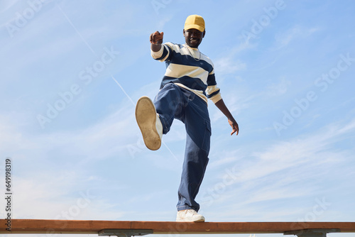 Minimal fashion shot of young black man wearing trendy outfit standing on roof against blue sky and kicking off to camera. photo