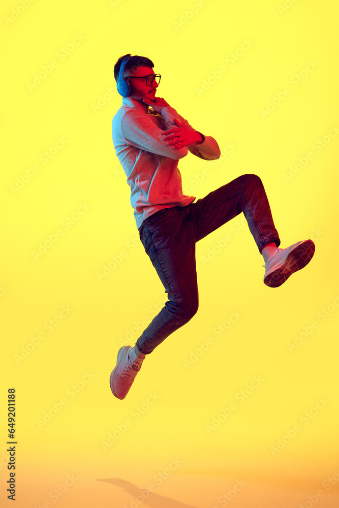 Full lenght portrait of thinking young guy listening music in headphones and jumping against on yellow background in red color light, filter.