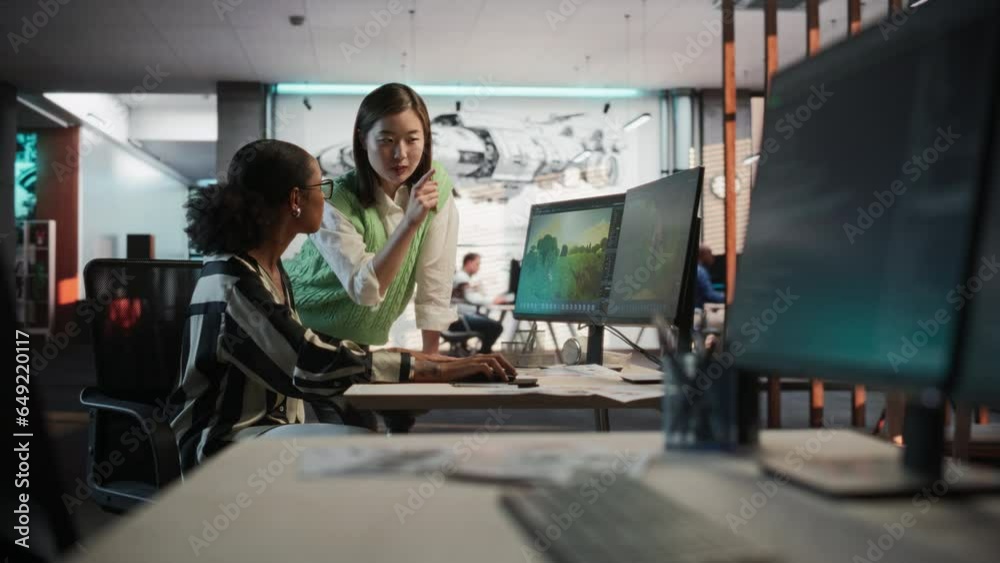 Black Female Game Designer Using Desktop Computer With 3D modelling Software And Talking to Asian Female Project Manager in Game Development Studio Office. Diverse Colleagues Discussing New RPG Game. Black Female Game Designer Using Desktop Computer With 3D modelling Software And Talking to Asian Female Project Manager in Game Development Studio Office. Diverse Colleagues Discussing New RPG Game.