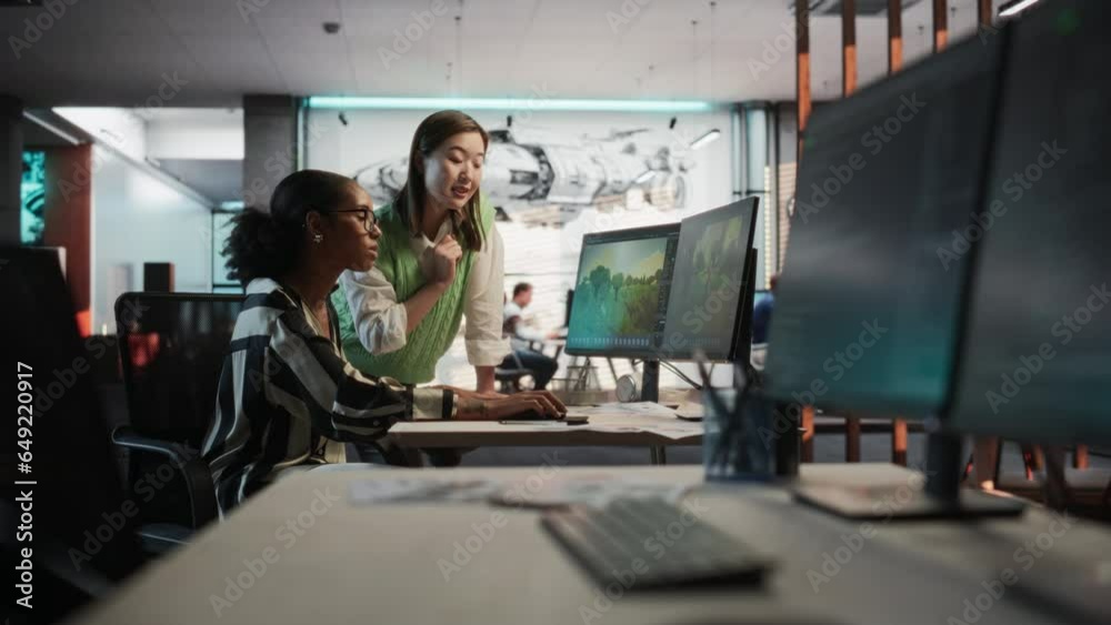Black Female Game Developer Using Desktop Computer With 3D modelling Software And Talking to Asian Female Project Manager in Game Design Studio Office. Two Diverse Colleagues Discussing New RPG Game. Black Female Game Developer Using Desktop Computer With 3D modelling Software And Talking to Asian Female Project Manager in Game Design Studio Office. Two Diverse Colleagues Discussing New RPG Game.