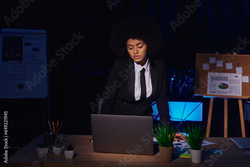 african american businesswoman in formal wear looking at laptop while working in office at night