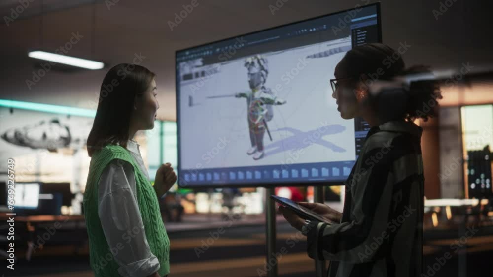 Black Female Game Designer Talking to Asian Female Project Manager And Standing Next To TV Set With 3D Game Character On Screen. Diverse Colleagues Discussing New RPG Game in Office. Slow Motion. Black Female Game Designer Talking to Asian Female Project Manager And Standing Next To TV Set With 3D Game Character On Screen. Diverse Colleagues Discussing New RPG Game in Office. Slow Motion.