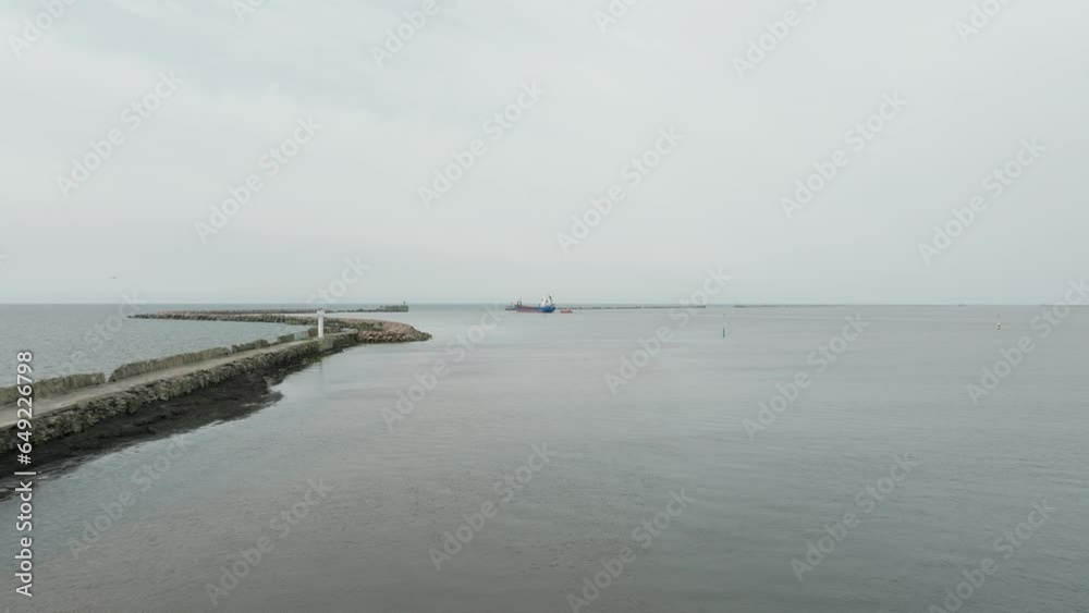Aerial establishing view of large blue cargo ship leaving Port of Liepaja (Latvia), Karosta bridge, slight overcast day, stone port pier, calm Baltic sea, distant wide drone shot moving forward low Aerial establishing view of large blue cargo ship leaving Port of Liepaja (Latvia), Karosta bridge, slight overcast day, stone port pier, calm Baltic sea, distant wide drone shot moving forward low