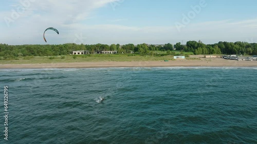Establishing aerial view of a group of people engaged in kitesurfing, sunny summer day, high waves, extreme sport, Baltic Sea Karosta beach (Liepaja), wide drone shot moving backward