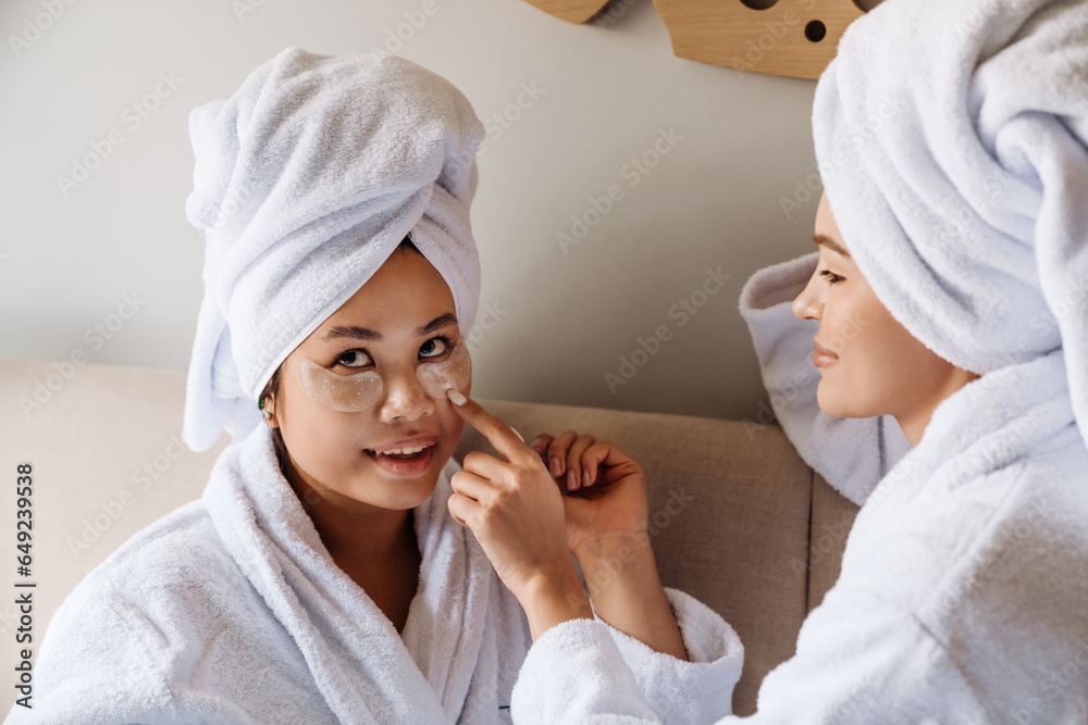 Two women wearing bathrobes and wrapped in towels using eye patches while sitting on bed Two women wearing bathrobes and wrapped in towels using eye patches while sitting on bed
