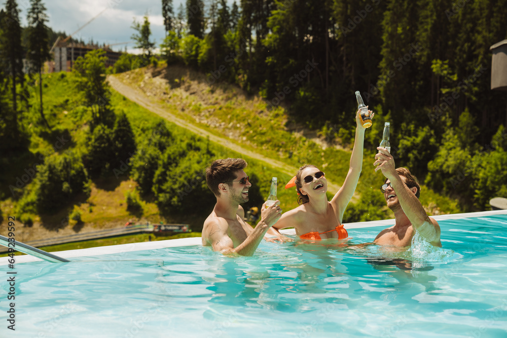 Group of cheerful friends relaxing in pool and drinking beer during vacation in mountains