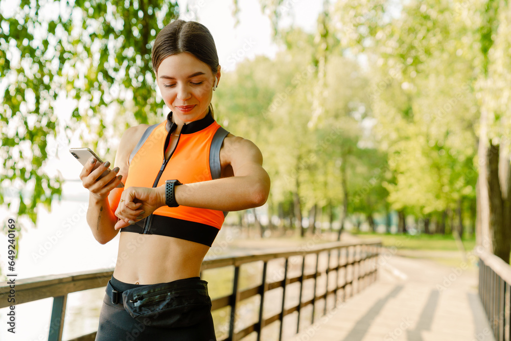 Smiling sports woman using mobile phone and checking time while standing in park