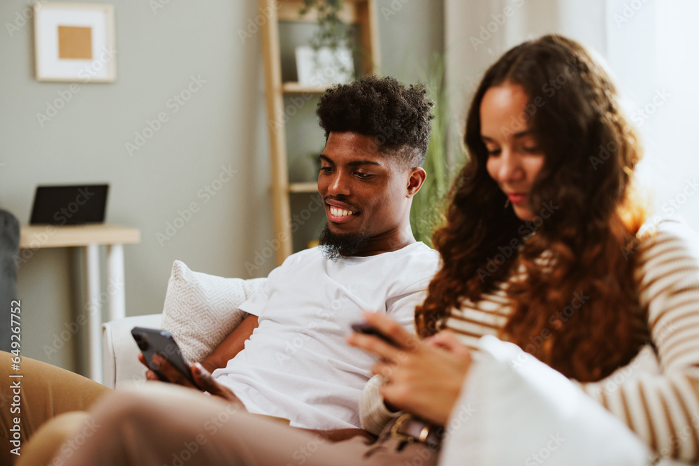 Obraz premium Young mixed race couple is sitting on a sofa and using a smart phone