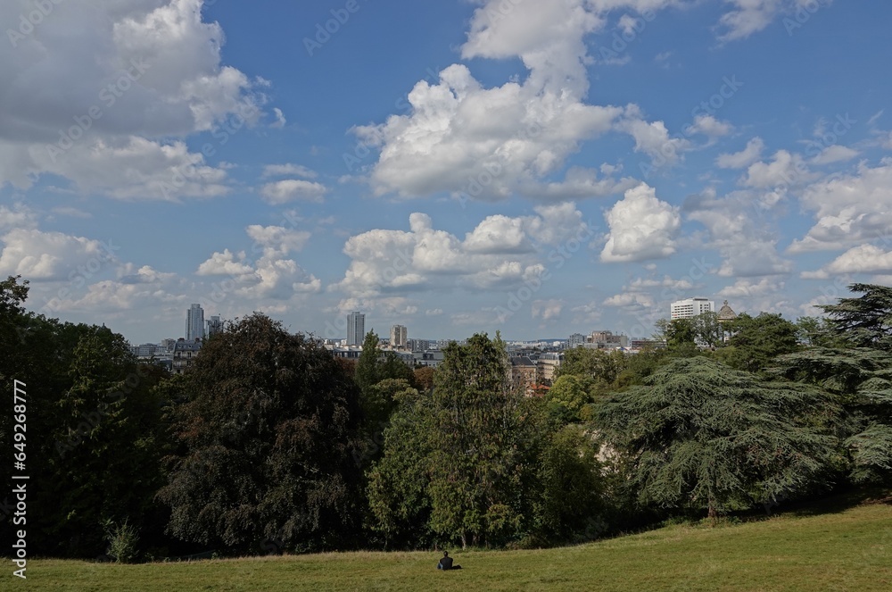 Vue du parc des Buttes-Chaumont sur Paris avec ciel bleu gros nuages blancs et une personne seule assise sur l'herbe