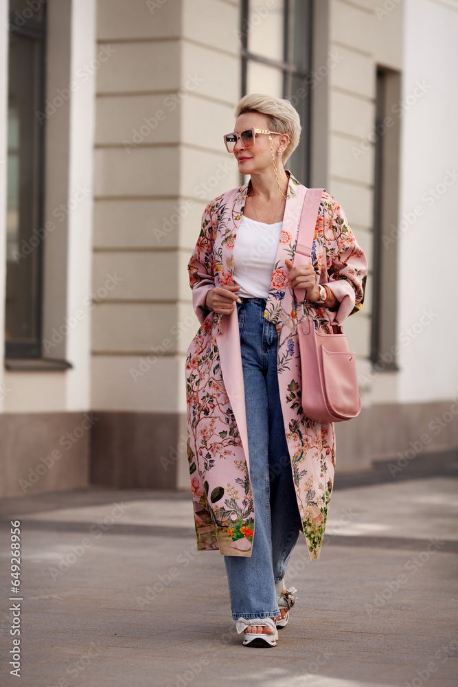 Beautiful Mature woman walking in city, wearing cargo jeans, pink floral coat, glasses, carrying bag. Urban street style, unique look of middle aged female model with short blonde hair