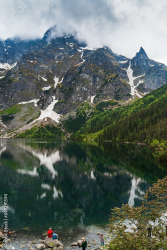 Fototapeta Naklejka Na Ścianę i Meble -  A serene, cloudy mountain landscape with reflections in Morskie Oko lake and tiny tourist figures on the shore. Iconic spot in Polish Tatra National Park for nature, travel, and adventure themes